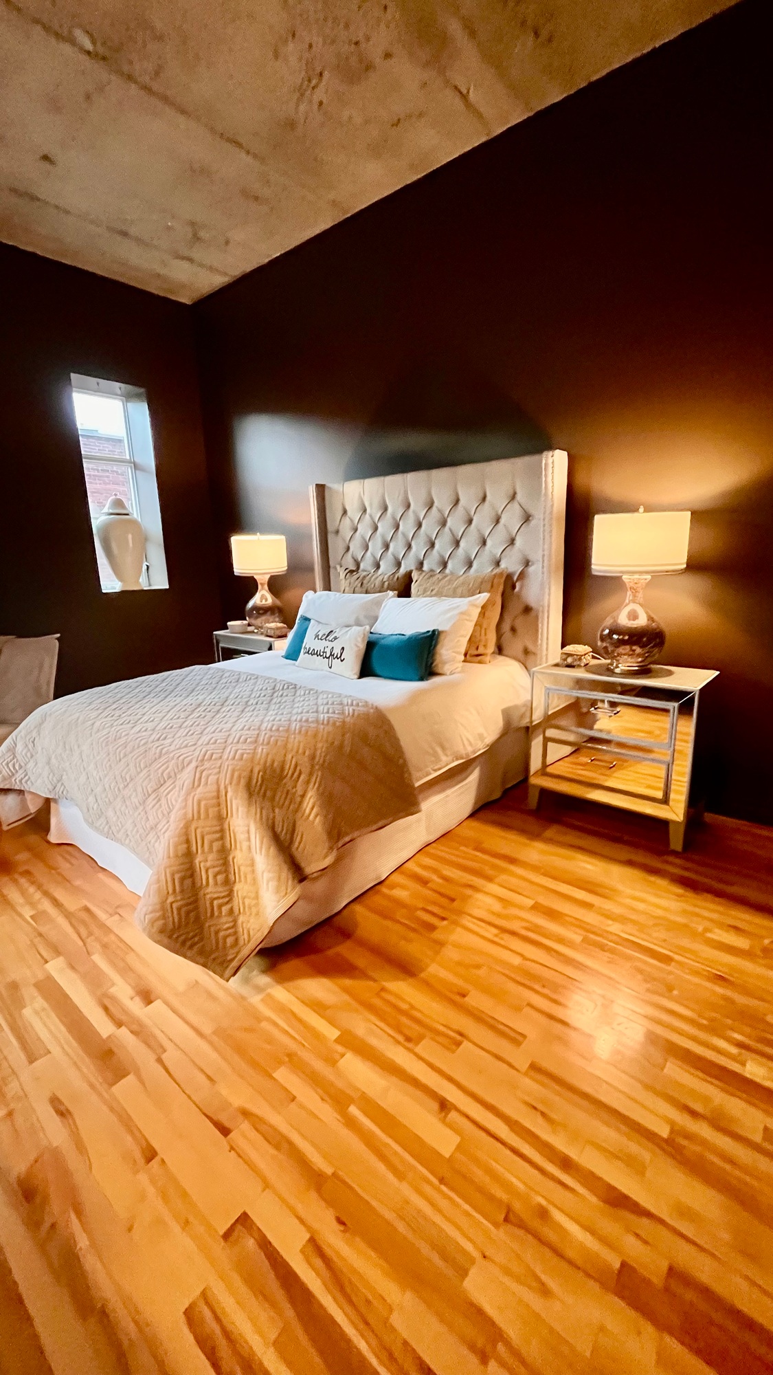 A loft style bedroom with grey upholstered headboard And mirrored night stands against a chocolate